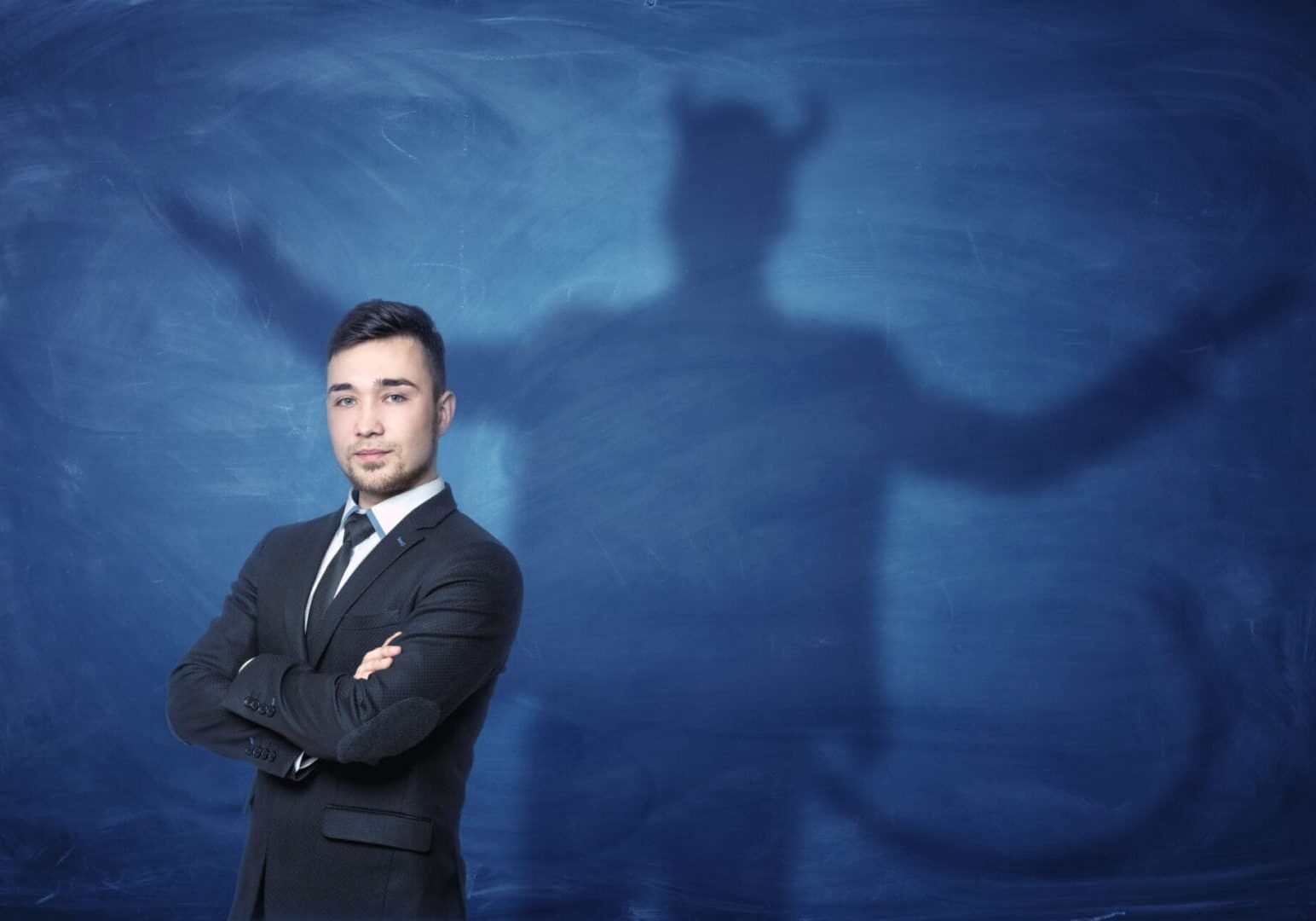 A businessman standing with his hands across and his shadow on a blue empty blackboard behind him with hands overspread, horns and a tail like a devil has. Self-confident behavior. Hidden feelings. Inner anger.